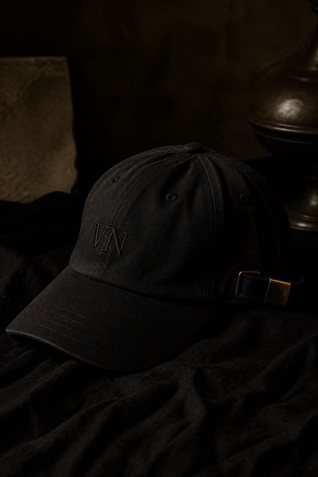 Black dad hat on dark linen, antique buckle catching a clay-gold light