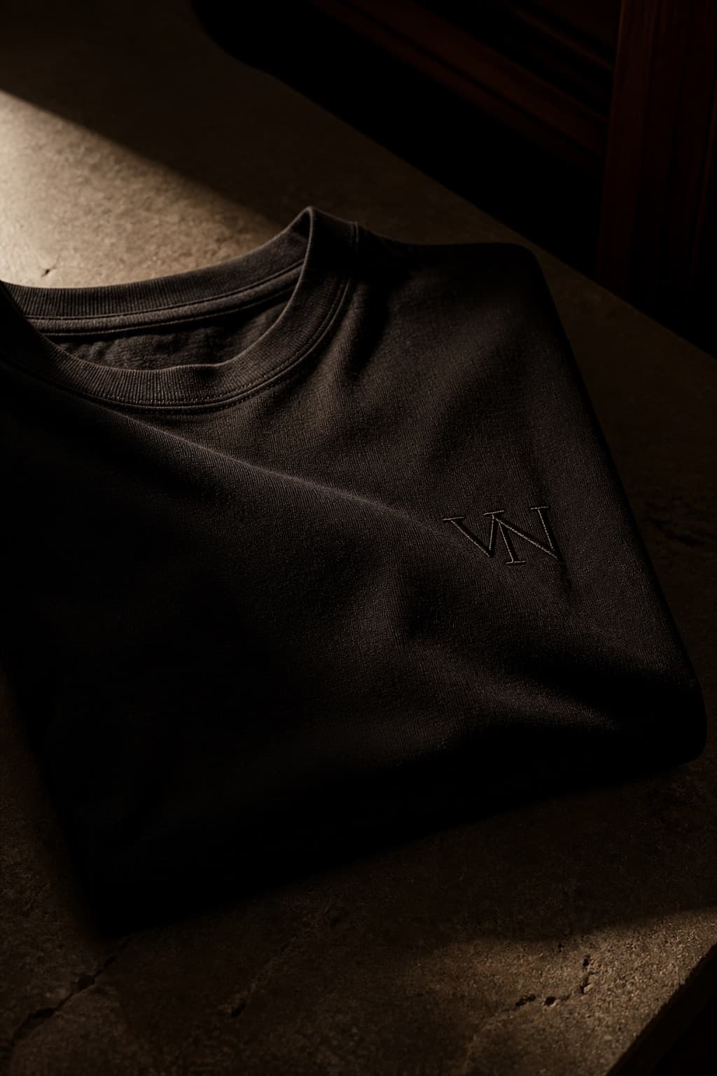 Black garment-dyed t-shirt draped on dark limestone, one crease catching a warm light